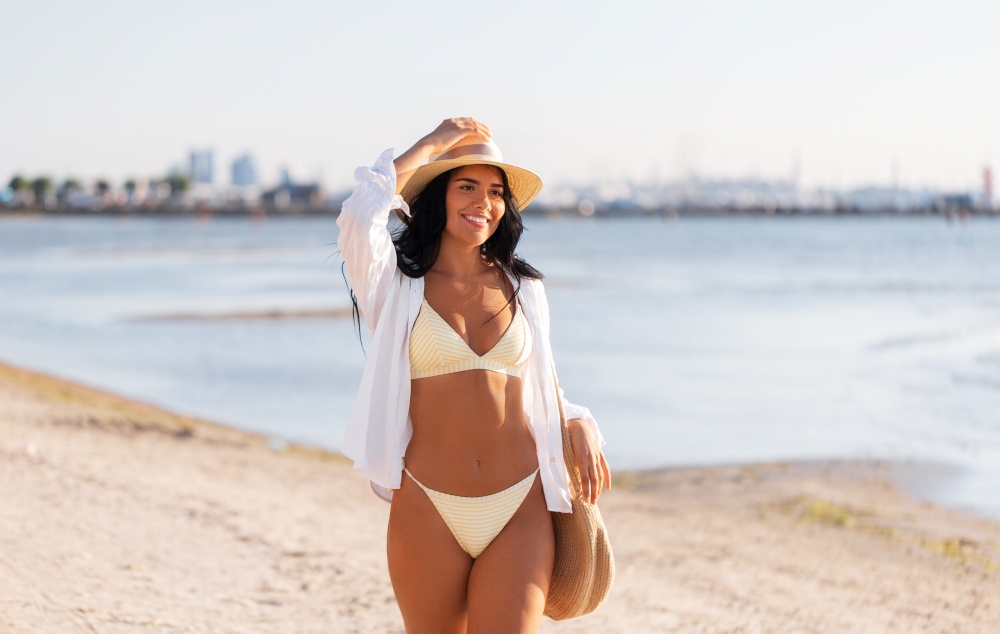 people, summer holidays and leisure concept - happy young woman in bikini swimsuit, white shirt and straw hat with bag walking along beach. happy woman in bikini and shirt walking on beach