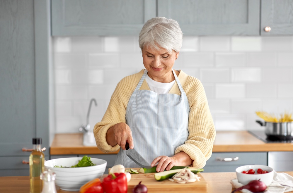 healthy eating, food cooking and culinary concept - happy smiling senior woman with knife chopping zucchini on kitchen at home. happy woman chopping zucchini on kitchen