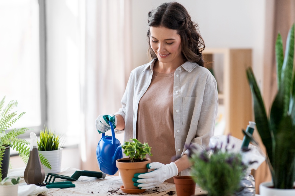 people, gardening and housework concept - happy woman in gloves with watering can planting pot flowers at home. happy woman planting pot flowers at home