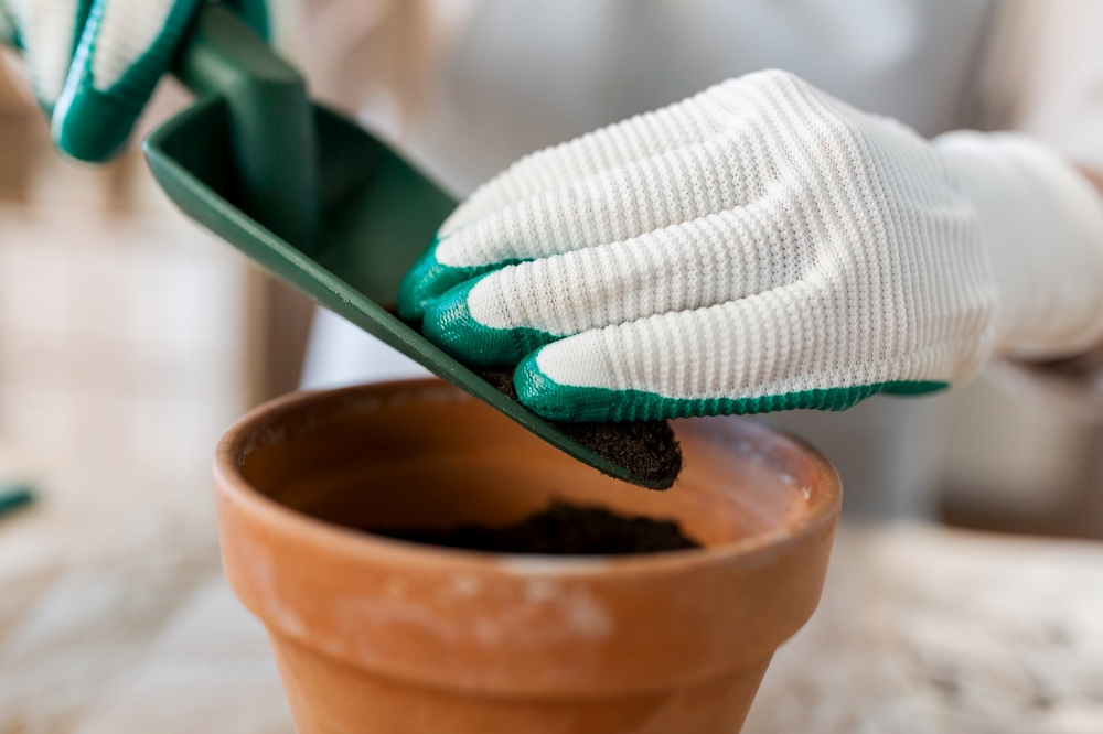 people, gardening and planting concept - close up of woman in gloves with trowel pouring soil to flower pot at home. close up of woman planting pot flowers at home