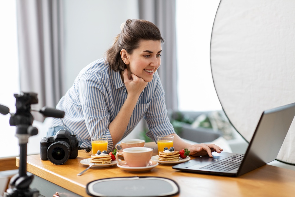 blogging, photographing and people concept - happy smiling female food blogger or photographer with laptop and camera in kitchen at home. female food photographer with laptop in kitchen
