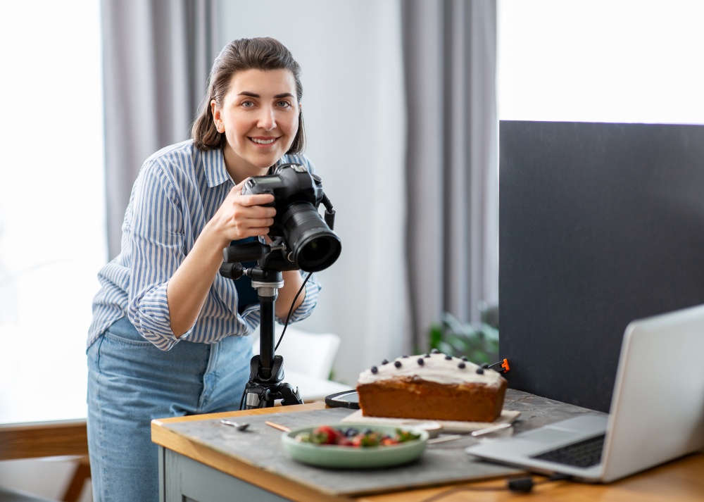 blogging, profession and people concept - happy smiling female food photographer with camera photographing cake in kitchen at home. food photographer with camera working in kitchen