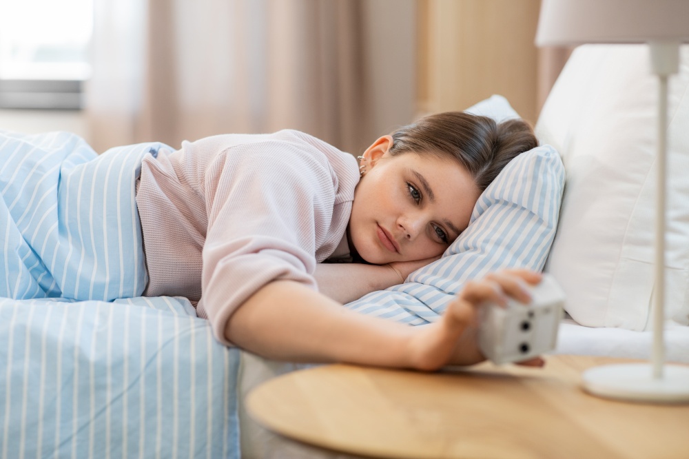 people, bedtime and rest concept - teenage girl looking at alarm clock lying in bed at home in morning. girl looking at alarm clock in bed in morning