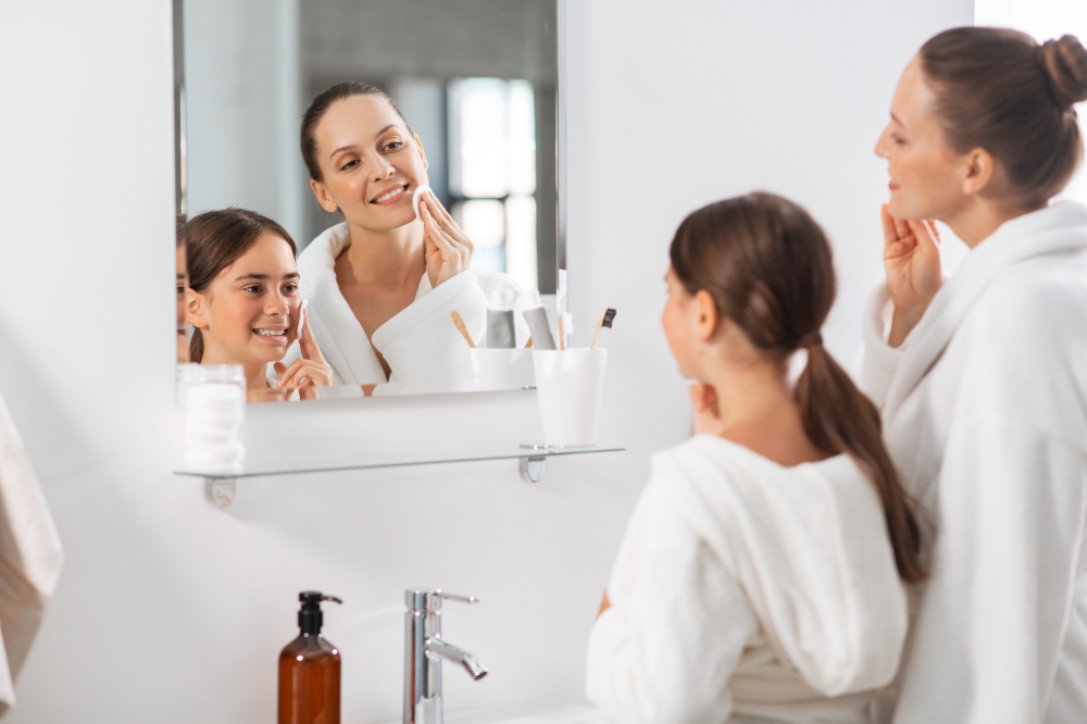 beauty, hygiene, morning and people concept - happy smiling mother and daughter with cotton pads cleaning face skin and looking to mirror at bathroom. mother and daughter with cotton pads cleaning skin