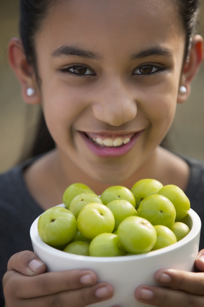 Close up of a smiling young girl holding a bowl full of indian gooseberries or amla.