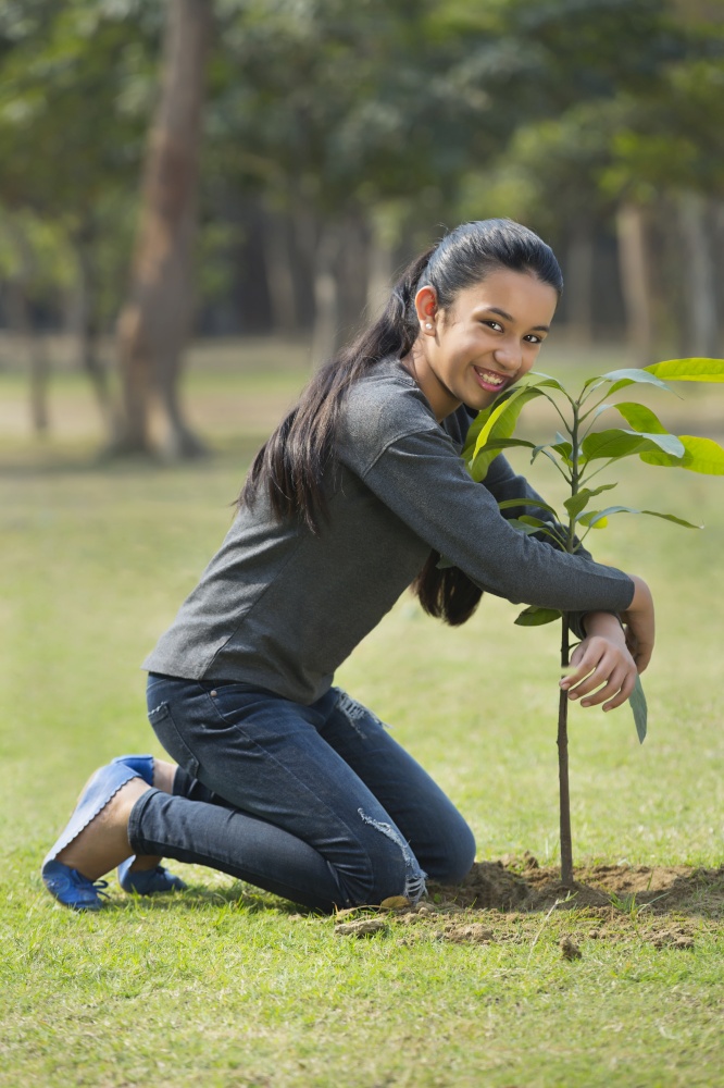 Happy young girl sitting on knees with her arms around a plant in park.