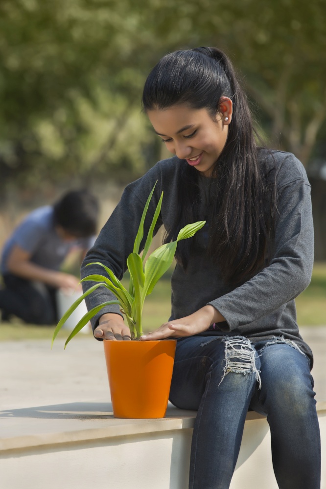 Young girl planting a small plant in a flower pot sitting in garden with a young boy sitting in the background.