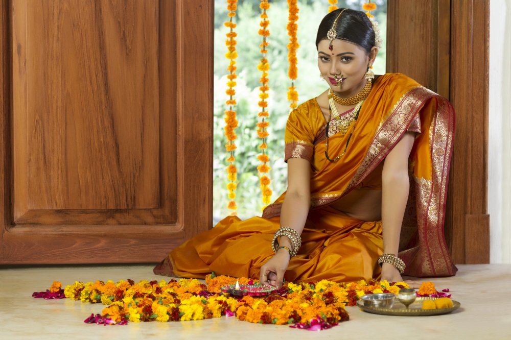 Woman dressed in traditional maharashtrian dress sitting near entrance with flower decorations pooja plate and diya on the floor.