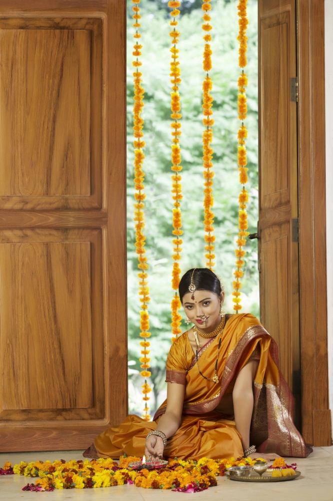 Woman dressed in traditional maharashtrian dress sitting near entrance with flower decorations pooja plate and diya on the floor.