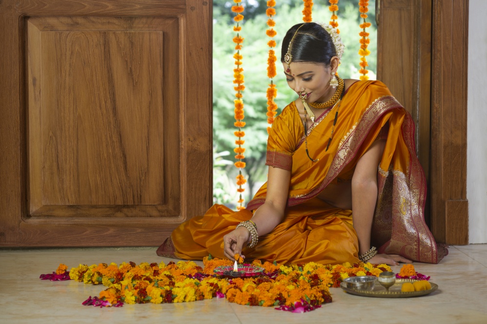 Woman dressed in traditional maharashtrian dress sitting near entrance with pooja plate diya and decorating floor with flowers.