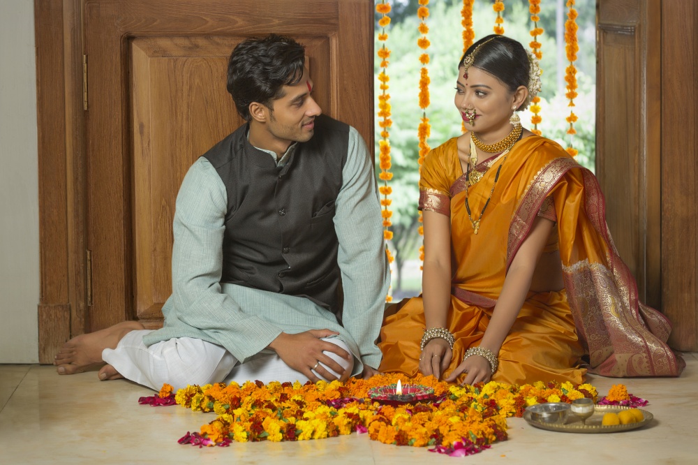 Happy maharashtrian couple in traditional dress sitting near entrance with flower decorations pooja plate and diya on the floor looking at each other.
