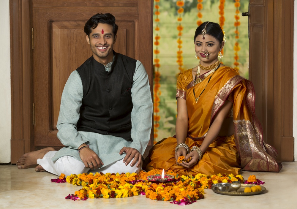 Happy maharashtrian couple in traditional dress sitting near entrance with flower decorations pooja plate and diya on the floor.
