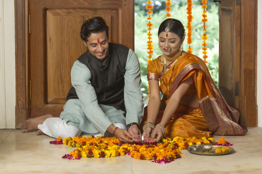 Happy maharashtrian couple in traditional dress sitting on the floor near entrance with flower decorations and pooja plate holding a diya.