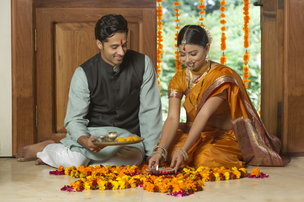 Happy maharashtrian couple in traditional dress sitting on the floor near entrance with flower decorations holding a diya and pooja plate.