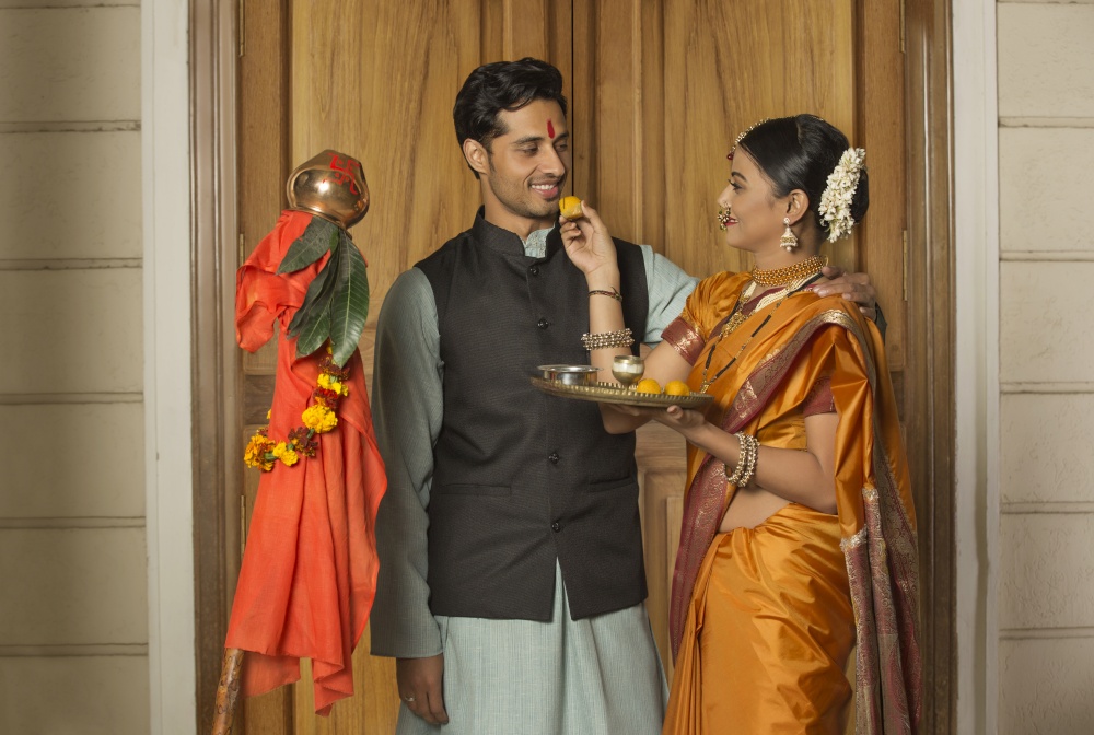Happy maharashtrian woman in traditional dress offering sweet to her husband from pooja plate while celebrating gudi padwa festival.