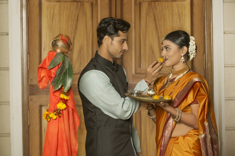 Maharashtrian man in traditional dress offering sweet to her wife while celebrating gudi padwa festival.