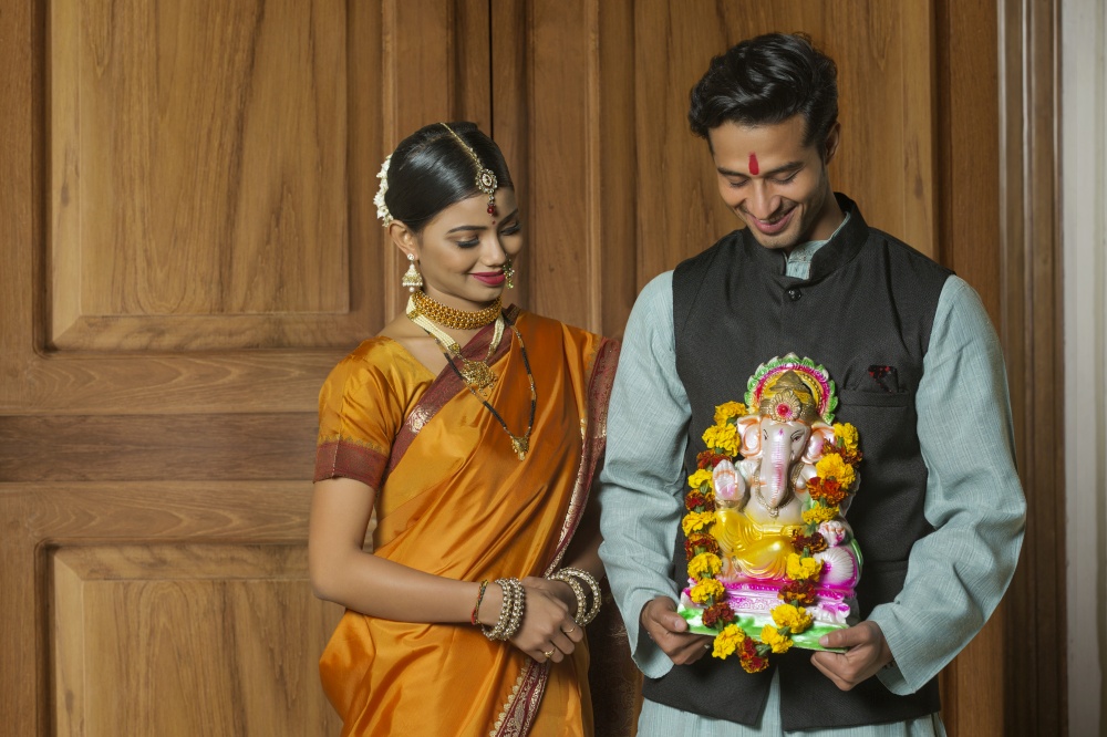 Maharashtrian couple in traditional dress celebrating ganapati festival holding a small statue of lord ganesha and looking at it.