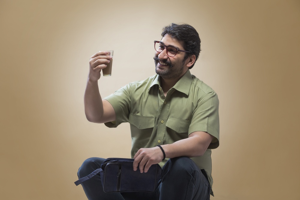 Smiling man wearing eyeglasses holding a small pouch in one hand and looking at a glass of tea in his other hand.