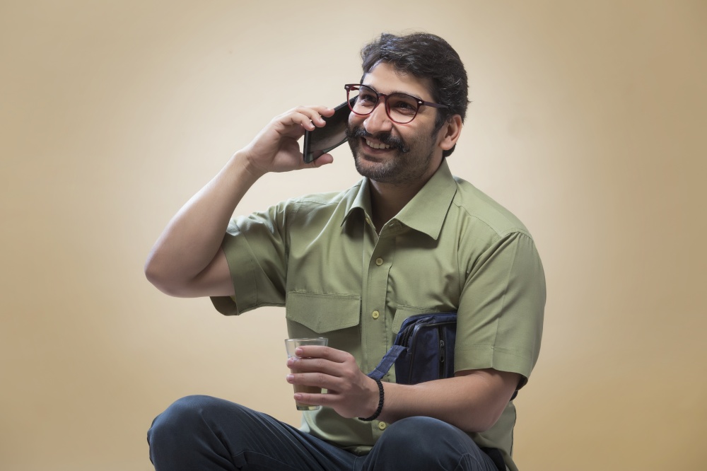 Smiling man wearing eyeglasses talking over mobile phone while holding a small pouch in one hand and a glass of tea in the other.