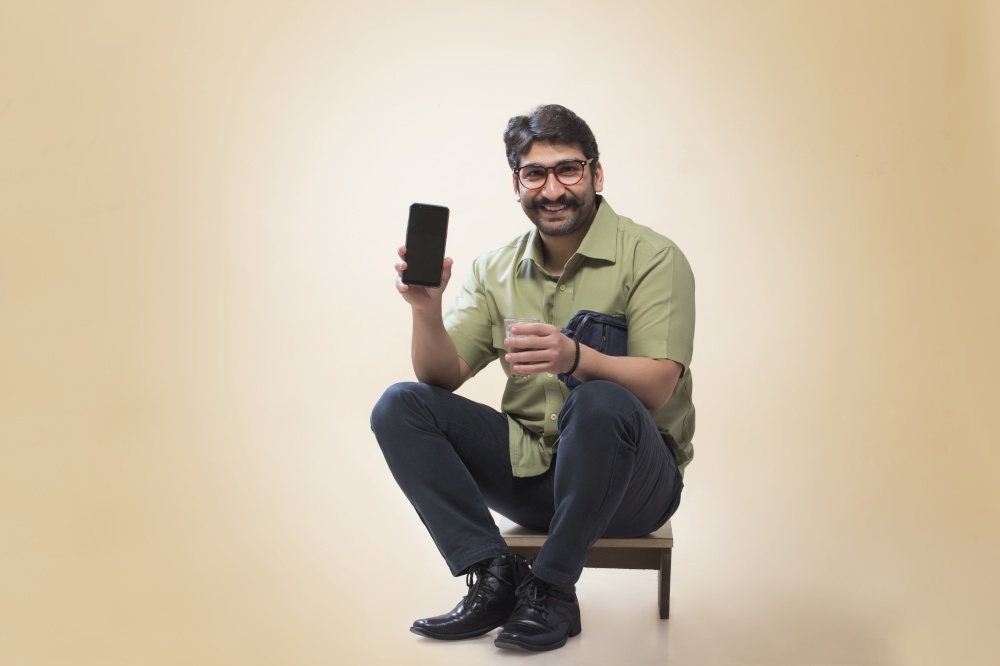 Smiling man wearing eyeglasses showing a mobile phone while holding a small pouch and a glass of tea.
