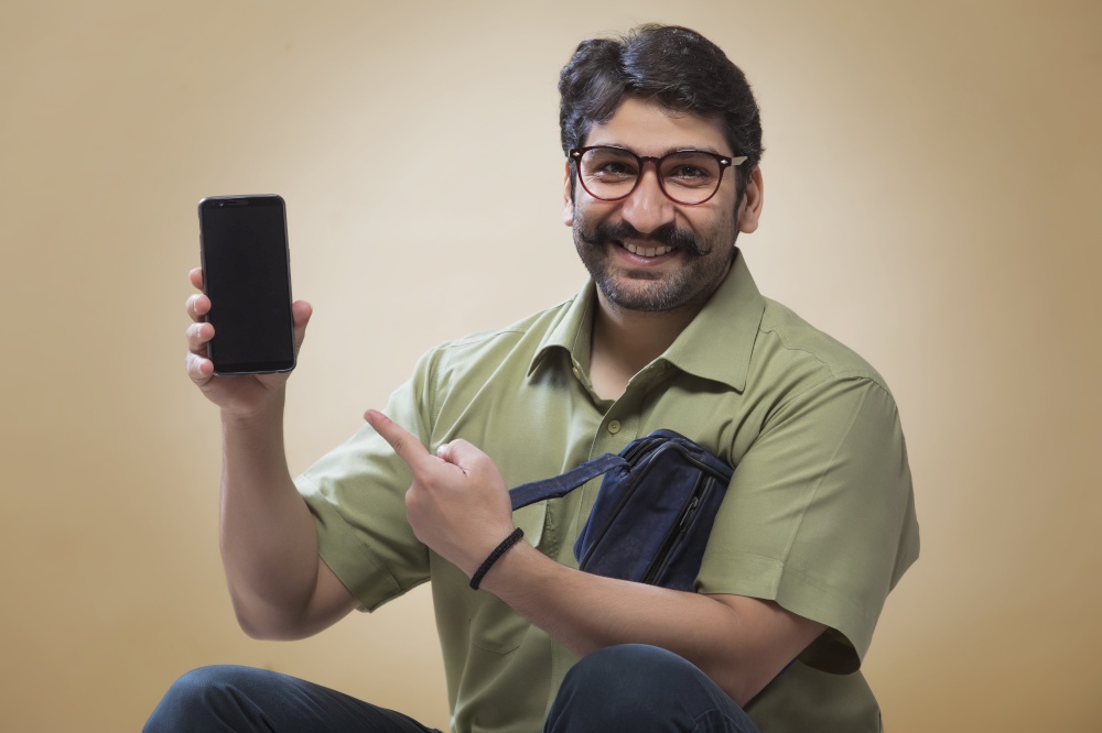 Smiling man wearing eyeglasses pointing towards a mobile phone which is held in other hand.