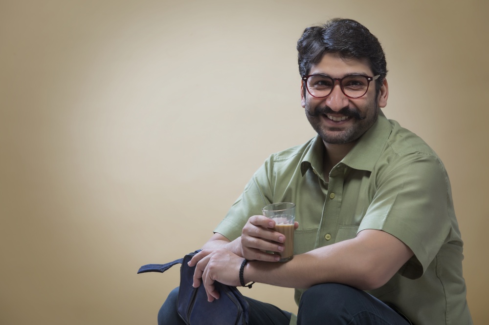 Smiling man wearing eyeglasses holding a small pouch in one hand and a glass of tea in the other.