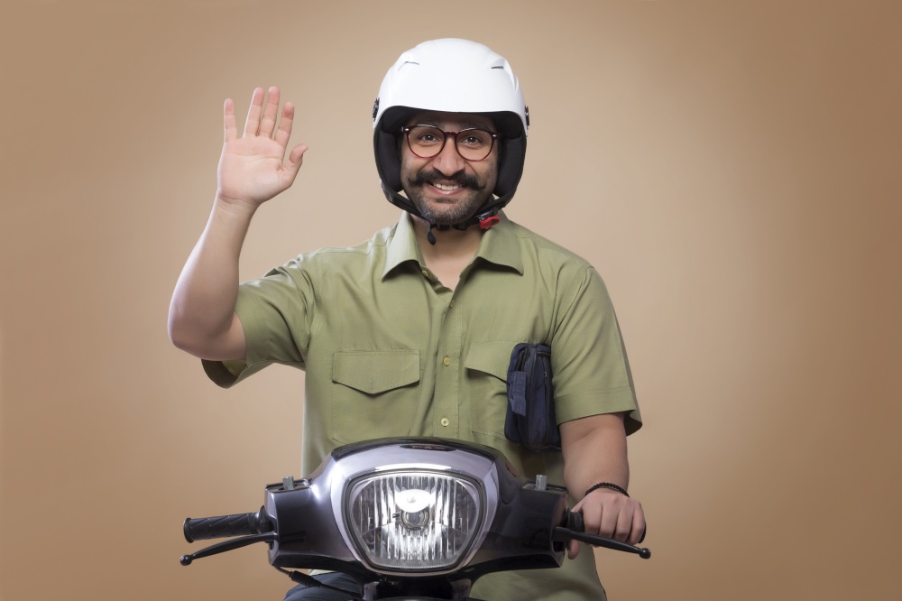 Smiling man with a small bag in his arms riding scooter wearing a helmet and showing palm.