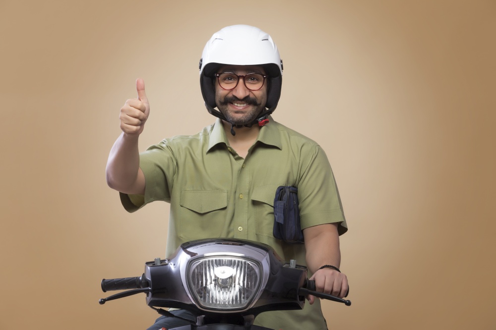 Smiling man with a small bag in his arms riding scooter wearing a helmet and showing thumbs up sign.