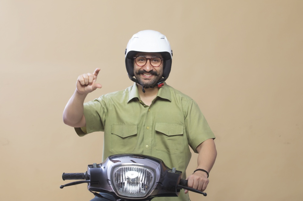 Man wearing helmet sitting on a scooter and showing gesture of holding a product with fingers.