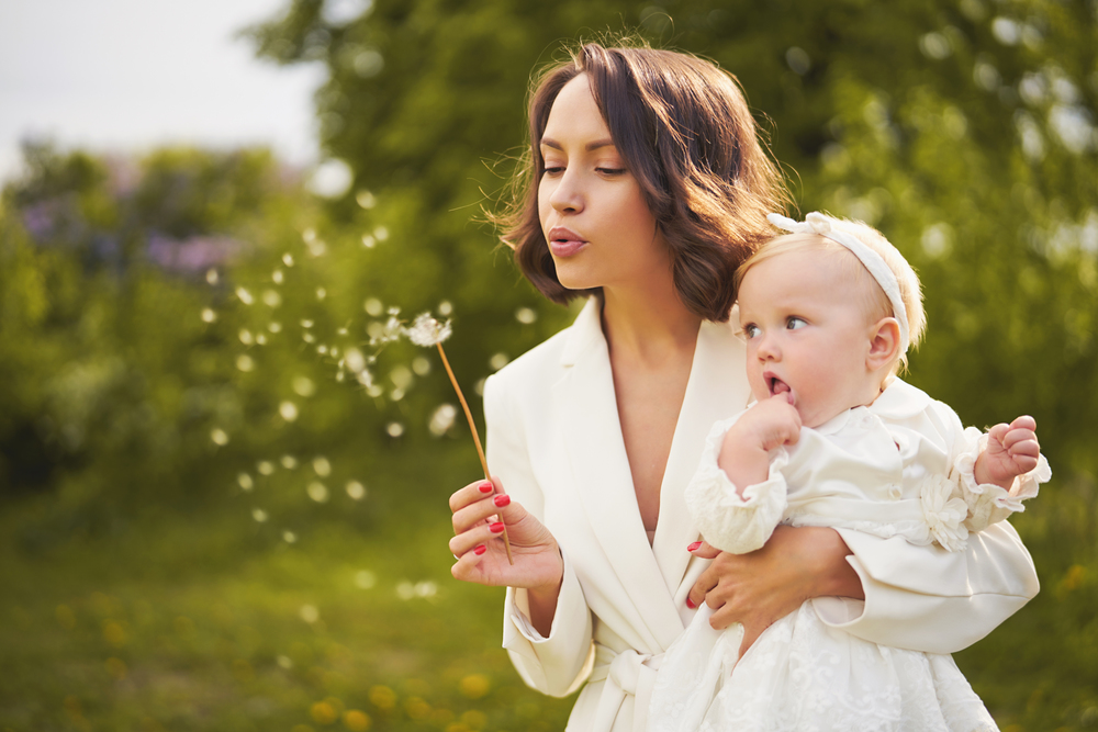 Outdoor fashion portrait of young beautiful mother and little cute daughter blowing to dandelion on green meadow. Summer image