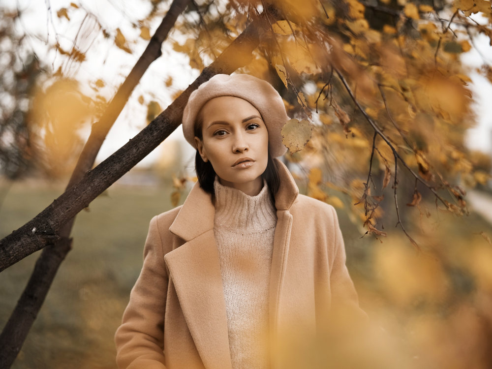 Outdoor fashion photo of young beautiful lady in beige coat, knite sweater and beret surrounded autumn leaves. Warm autumn