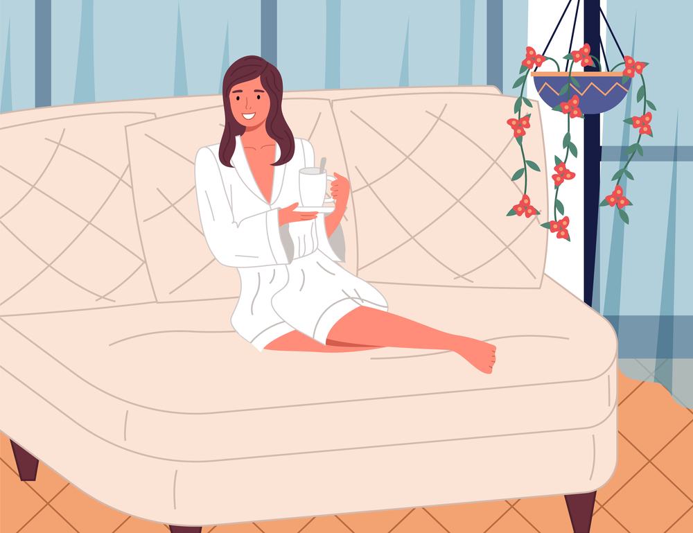 Young beautiful smiling woman sitting with bent legs on a large white comfortable sofa with cup of tea or drink, white bathrobe. Girl in the spa. Beauty treatments and relaxation. Cozy interior. Beautiful young girl in terry white bathrobe on the couch with cup of drink. Spa treatments