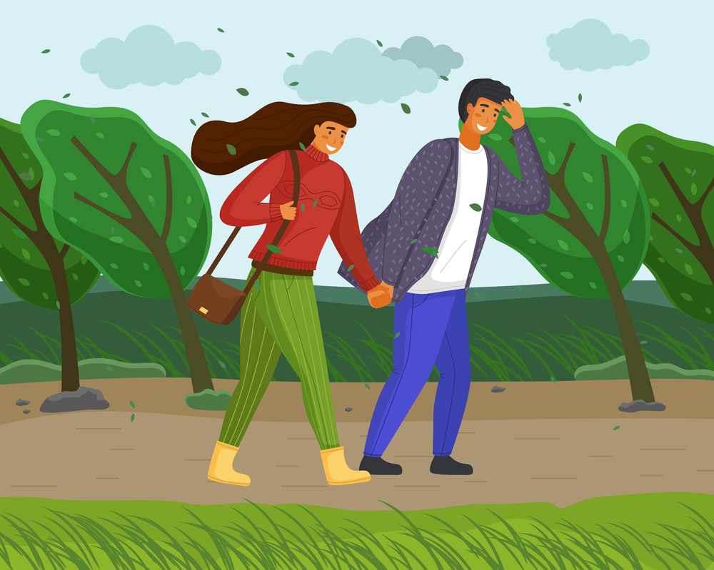 Woman and man in casual clothes walk in the park on the road along the tree alley in windy weather. Strong wind blows, trees bend to the ground, leaves are flying, young people smiling holding hands. Woman and man in casual clothes walk in the park on the road along the tree alley in windy weather