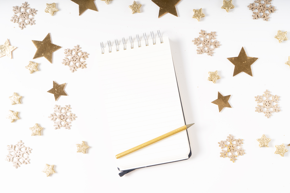 Wish list for Cristmas and New Year. Holiday decorations and ruled notebook with wish list on white desk, flat lay over white. Christmas flat lay scene with golden decorations