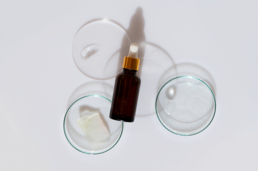 Glass petri dish with transparent pure serum for skin care with serum cosmetic bottle on white background, top view. Concept laboratory tests and research, making and testing cosmetic. Glass petri dish with transparent pure serum for skin care