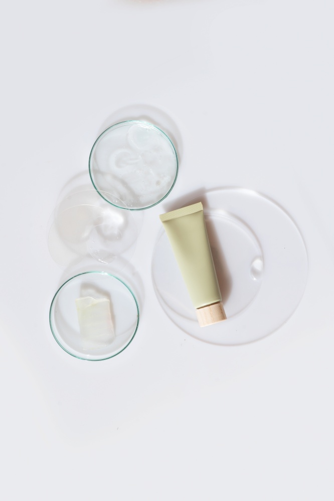 Glass petri dish with transparent pure serum for skin care with green cosmetic tube on white background, top view. Concept laboratory tests and research, making and testing cosmetic. Glass petri dish with transparent pure serum for skin care