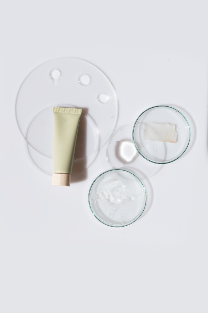 Glass petri dish with transparent pure serum for skin care with cosmetic tube on white background, top view. Concept laboratory tests and research, making and testing cosmetic. Glass petri dish with transparent pure serum for skin care
