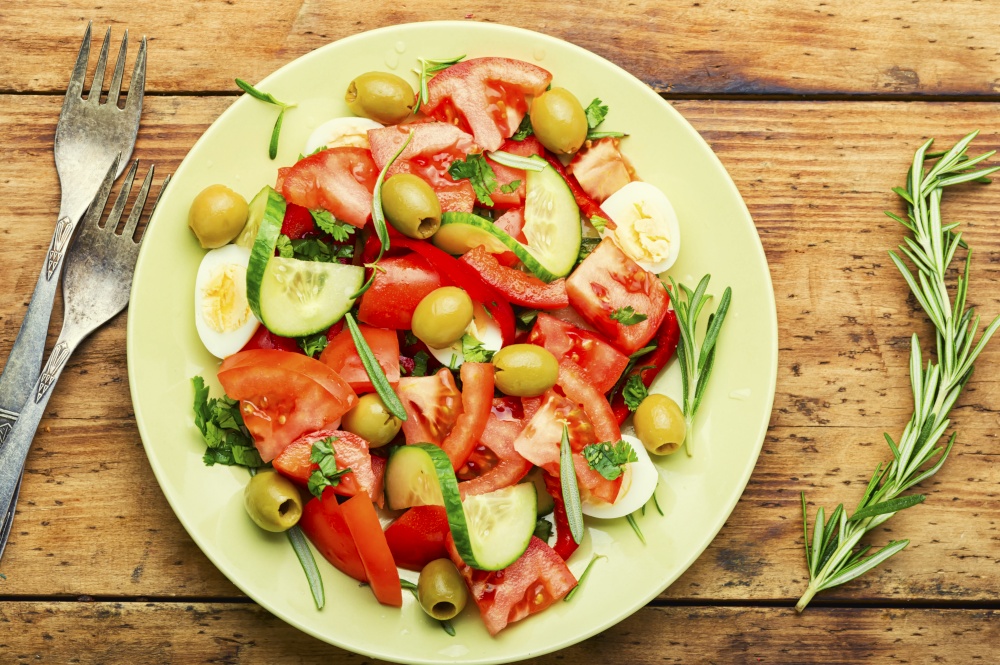 Vegetable salad of tomatoes, cucumbers, olives and eggs on wooden background. Salad with vegetables, olives, eggs and rosemary
