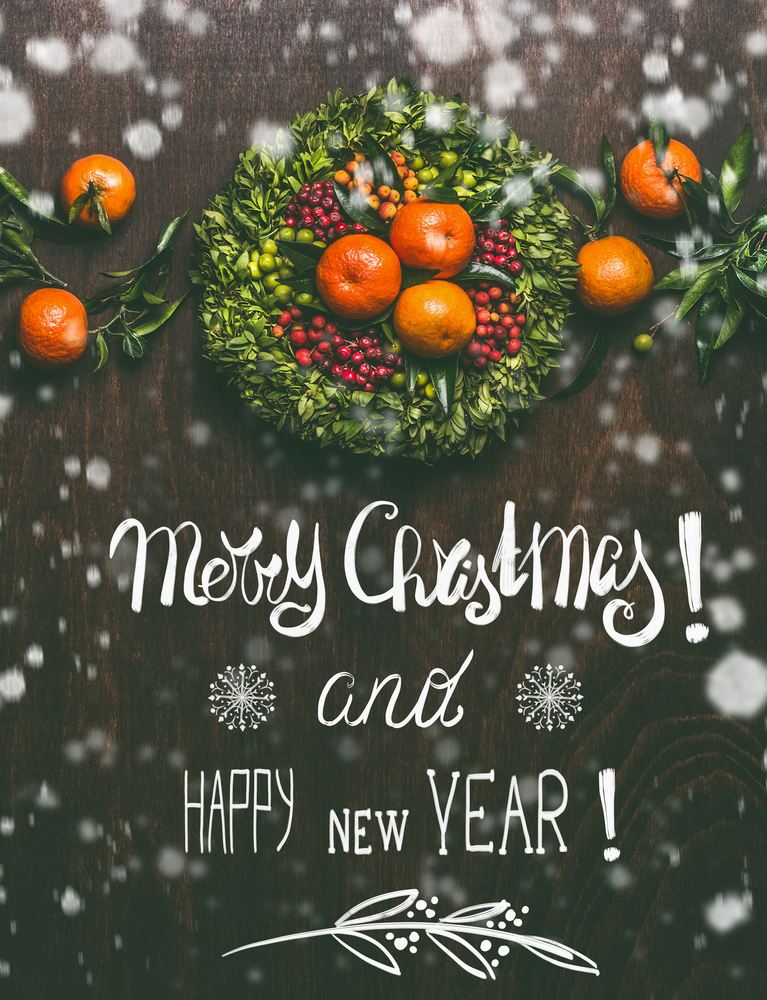 Merry Christmas and Happy New Year lettering greeting card with painted snow and tangerines garland with leaves wreath on dark rustic background, top view.