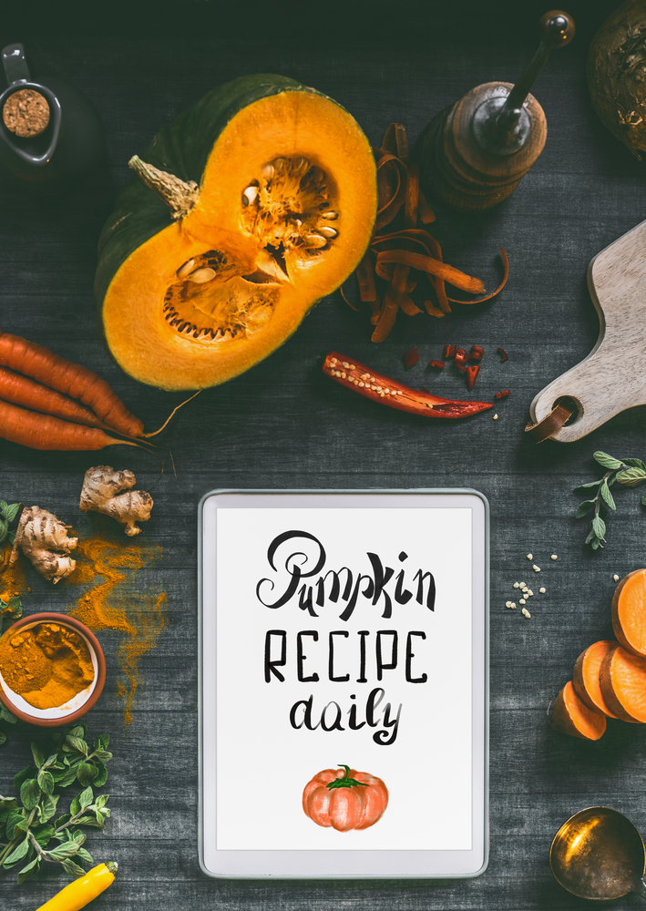 Tablet pc with text lettering: Pumpkin recipe daily. Orange cooking ingredients: sweet potato, carrots, turmeric powder,chili and ginger for tasty cuisine. Healthy and clean vegetarian food and eating