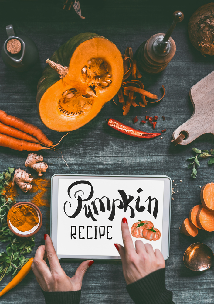 Female hand pointing with finger on tablet pc with text lettering: Pumpkin recipe on kitchen table background with orange cooking ingredients: sweet potato, carrots, turmeric powder,chili and ginger