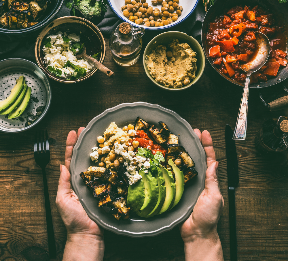 Female hands holding with healthy vegetarian bowl with various grilled vegetables, avocado and  chickpea hummus on rustic background, top view. Clean food and dieting nutrition concept