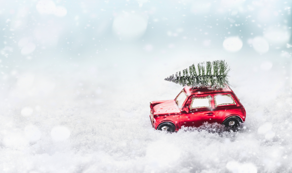 Christmas tree on roof of red toy retro car in snow through snowy winter wonder land with snowfall. Creative Christmas holiday concept. Copy space for your greeting and design. Front view