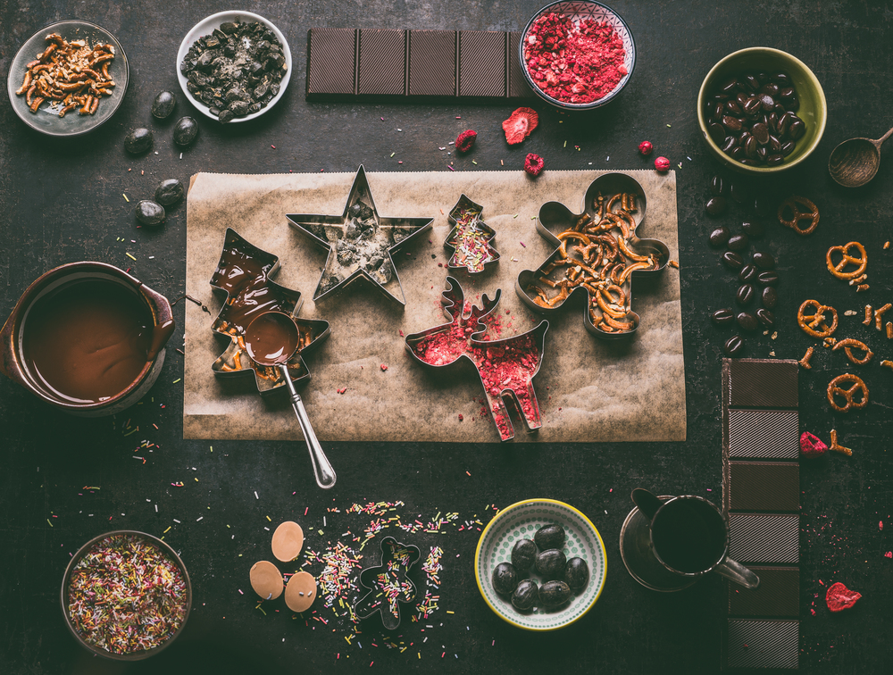 Homemade Christmas chocolate bars making. Christmas cutters with various toppings and flavorings. Melted chocolate in bowl with spoon on dark rustic kitchen table background, top view. Edible gifts