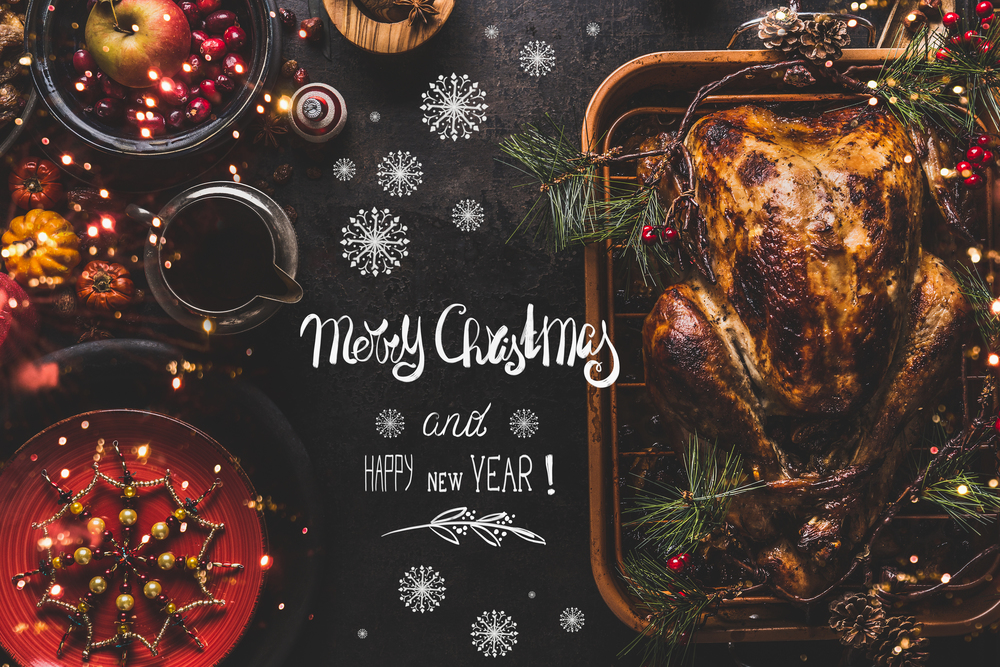 Merry Christmas and Happy New Year greeting text lettering on Christmas dinner table with whole roasted turkey served with sauce, red plates, cutlery, decoration and burning candles