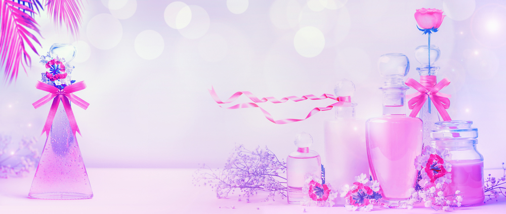 Various cosmetic product bottles in neon color with ribbons and flowers standing on pink purple background with bokeh. Skin care, cosmetic shop, sale and abstract beauty concept. Banner or template