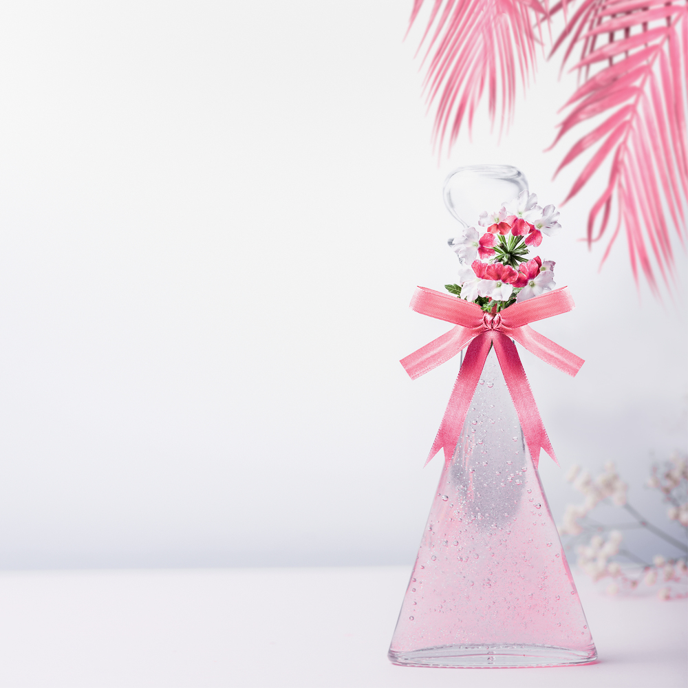 Stylized cosmetic glass bottle with pink ribbon and flowers and hanged palm leaves standing on white gray background with copy space. Skin care and beauty concept