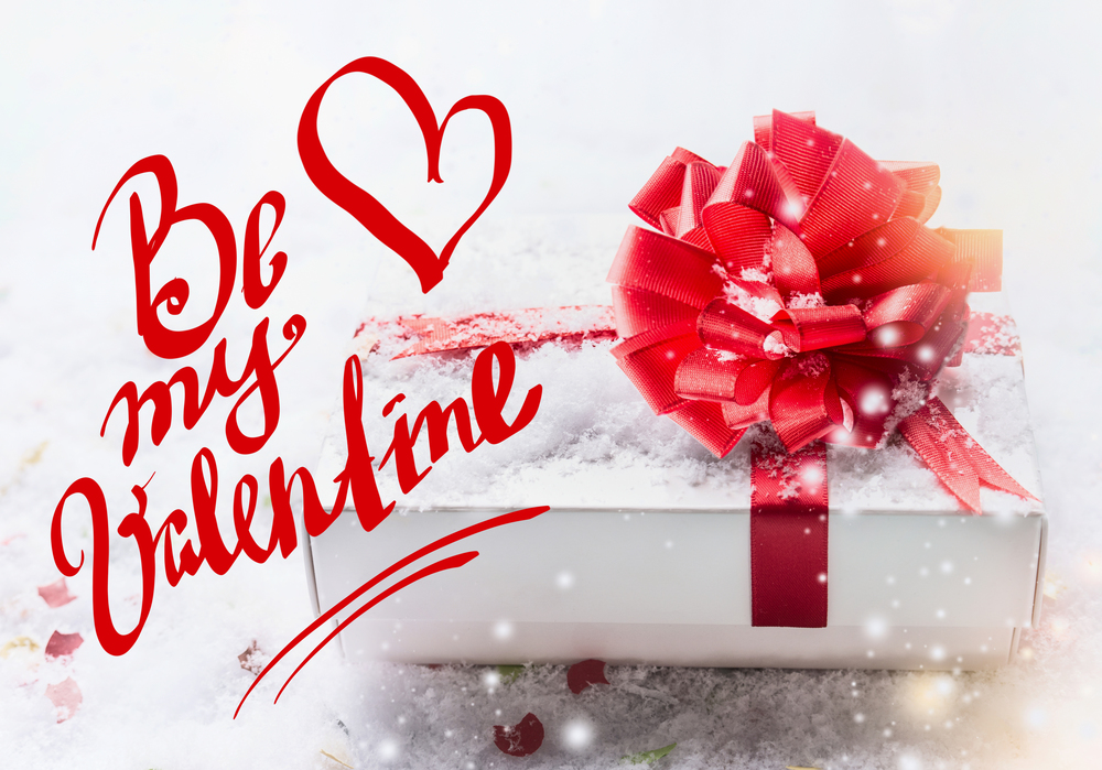 Be my Valentine text lettering with white gift box, red bow on snow with bokeh and snowfall. Valentines day, declaration of love concept