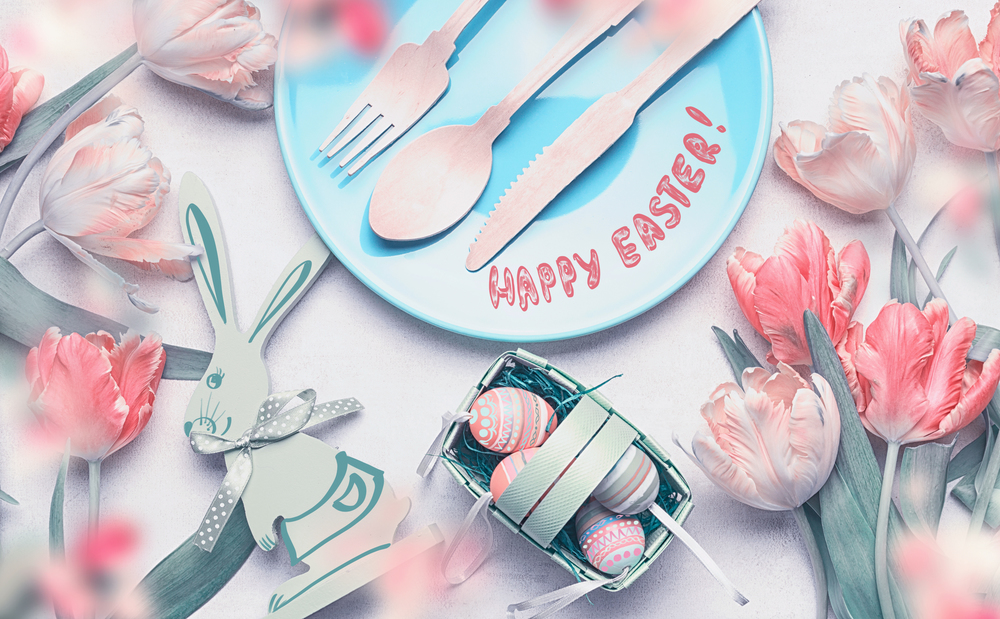 Happy Easter table setting with Easter eggs and decoration accessories, plate and cutlery, tulips flowers and funny rabbit on white background, top view, flat lay. Handwritten style text lettering