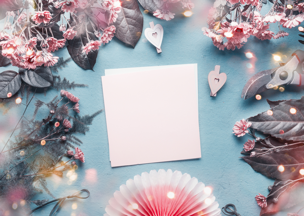 Blank greeting card on blue florist desk with pink flowers, hearts and paper origami fan. Abstract greeting concept. Mothers day, birthday mock up. Copy space. Top view, flat lay